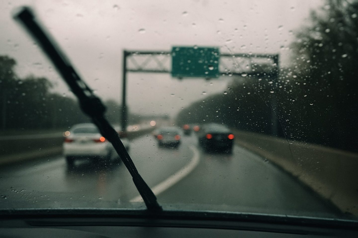 A rain-spotted windshield with a wiper mid-sweep, brake lights ahead on a curving interstate ramp under a green overhead sign.